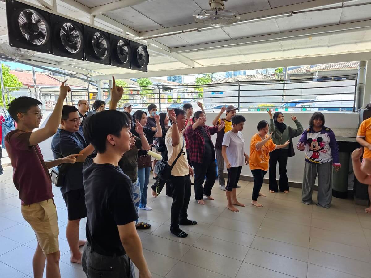 AGM Group volunteers and residents dancing with arms raised in the open hall of PKKII Insan Home in Happy Garden, Kuala Lumpur