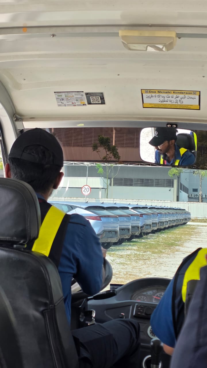 Perodua stockyard operations viewed from a shuttle vehicle, showing a long row of parked finished units and a 20 km/h yard speed limit sign